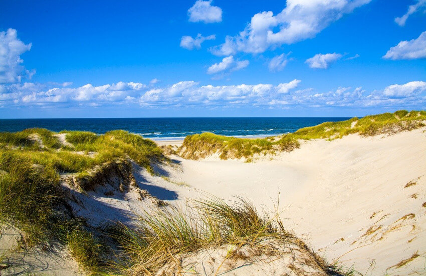 Ein weißer unebener Sandstrand teilweise mit Dünen bewachsen und dahinter das Meer.