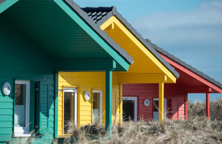 Ein grünes, ein gelbes und ein rotes Ferienhaus aus Holz, etwas versetzt zwischen Dünen.