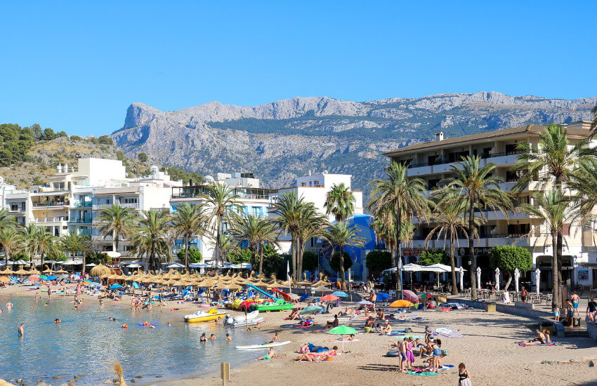 Ein Sandstrand mit vielen Badegästen, dahinter Hotels mit dem Gebirge im Hintergrund.