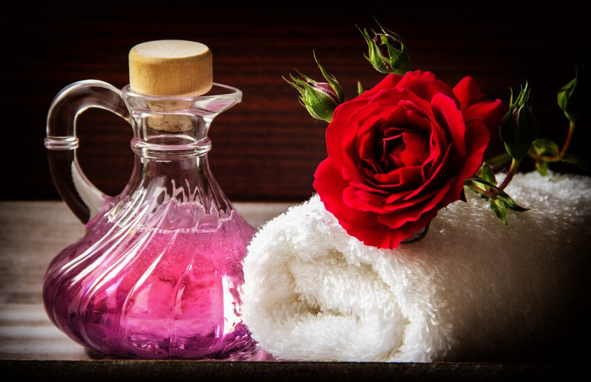 Eine Wellness Karaffe mit einem lila farbenen Duft und ein daneben ein zusammengerolltes Handtuch mit einer roten Rose oben drauf.