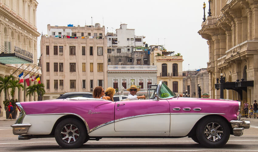 Ein weißpinkes Oldtimer Cabrio mit zwei Touristinnen in der Altstadt von Havanna.