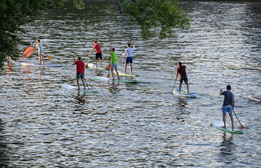 Sieben Personen Paddeln stehend mit einem Stand-Up-Paddle Board auf einem See.