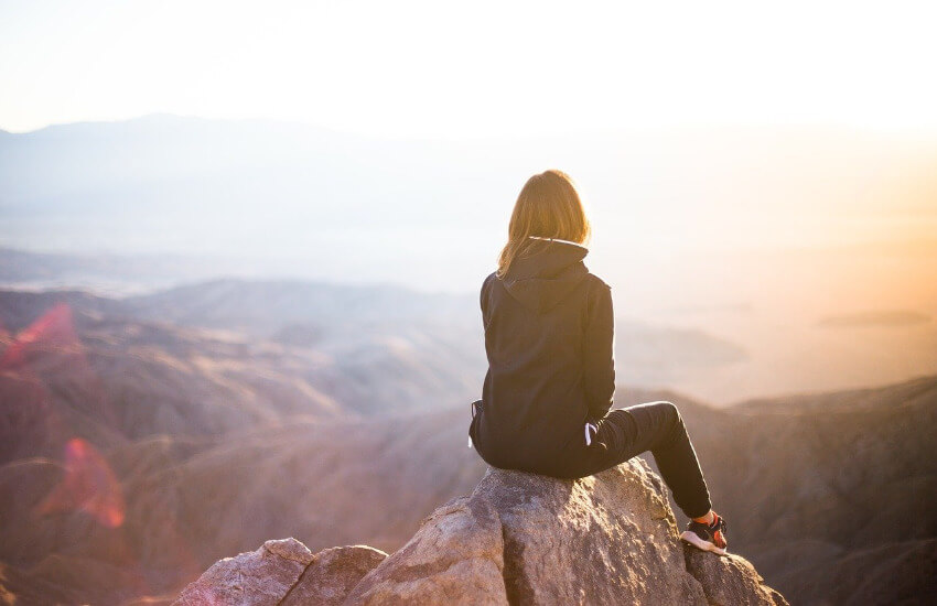 Eine Frau sitzt auf einem Fels oben am Berg und blickt Richtung Sonnenaufgang.