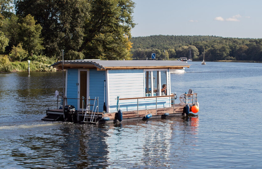 Ein Hausboot mit hellblauer Holzverschalung unterwegs auf einem See.