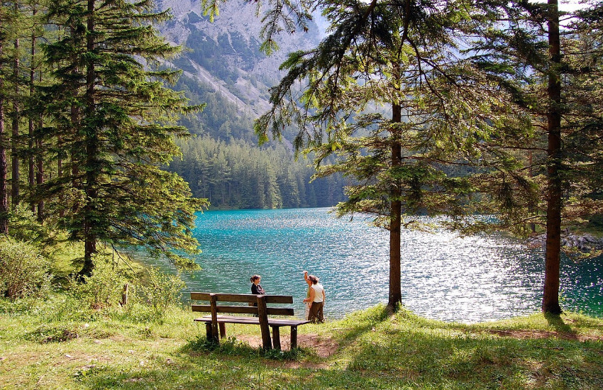 Ein See mit türkisem Wasser, umgeben von Wald und Bergen und einem schmalen Einstieg mit einer Sitzbank davor.