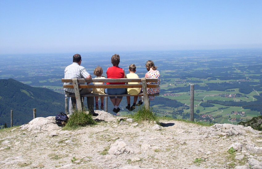 Eine Familie mit drei Kindern sitz auf einer Bank auf einem Berg und genießt die Aussicht auf die grüne Landschaft.