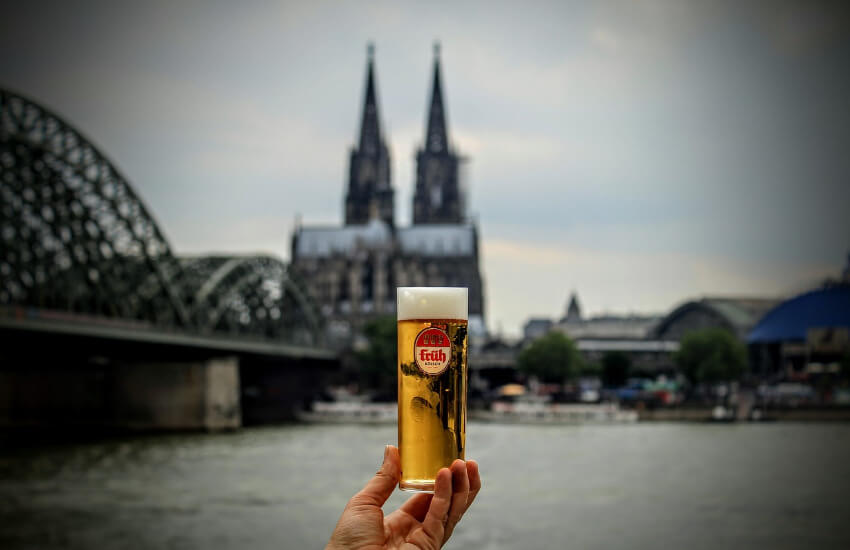 Eine Hand hält ein Kölsch in die Luft, wobei im Hintergrund der Kölner Dom zu sehen ist.