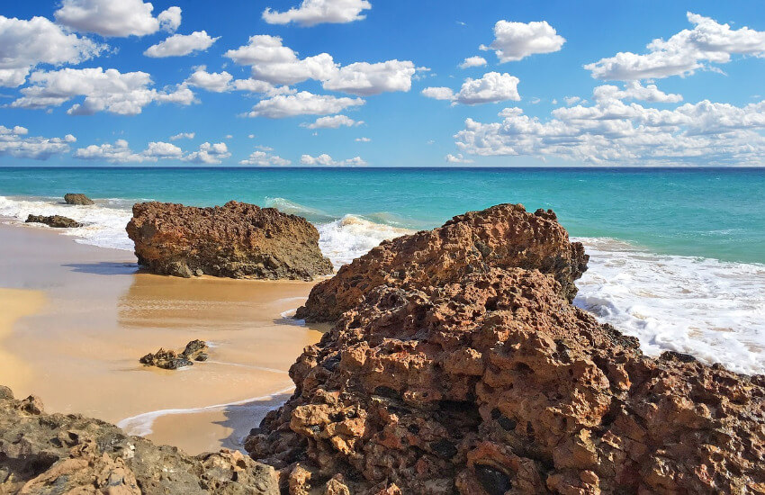 Türkises Meer und ein Sandstrand mit vereinzelt großen Steinen dazwischen.