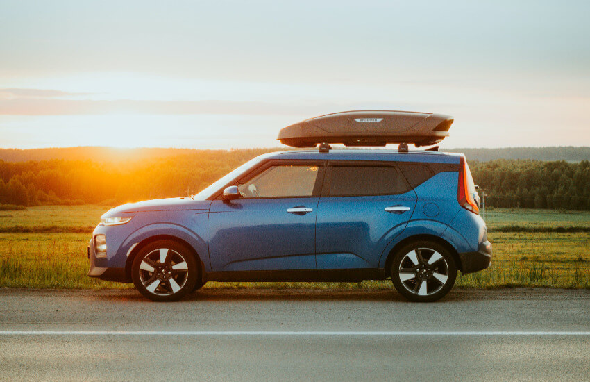 Ein blaues Auto mit einer schwarzen Dachbox steht am Straßenrand und im Hintergrund ein Sonnenaufgang.