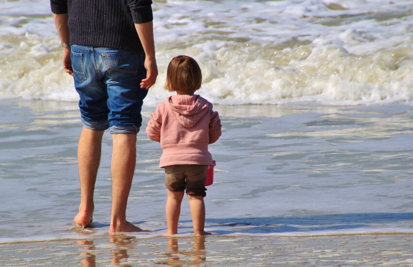 Ein Vater und ein Kind mit hochgezogenem Hosenbein stehen in sanften Wellen der Ostsee.