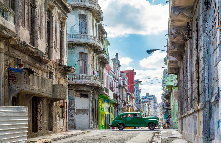 Grüner Oldtimer fährt durch eine von bröckelnden, bunten Kolonialbauten gesäumte Gasse in Havanna.