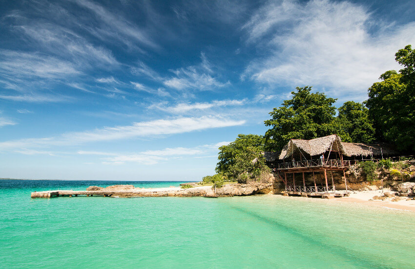 Holzhütte mit Strohdach am Strand Kubas. Türkisblaues Wasser und felsige Küste unter blauem Himmel.